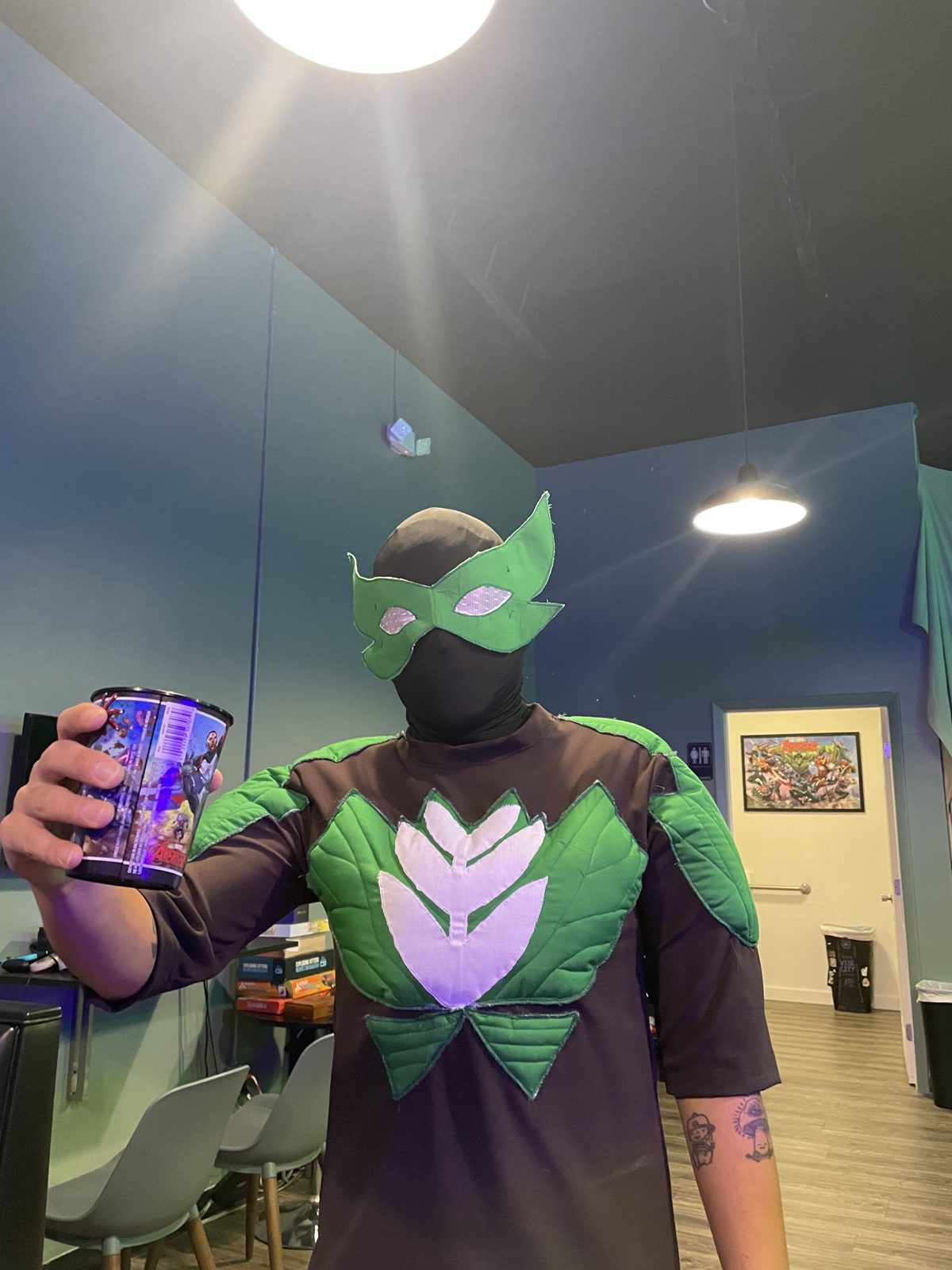 Komic Kava regular in a leaf-styled superhero costume holding a comic-print cup at the bar